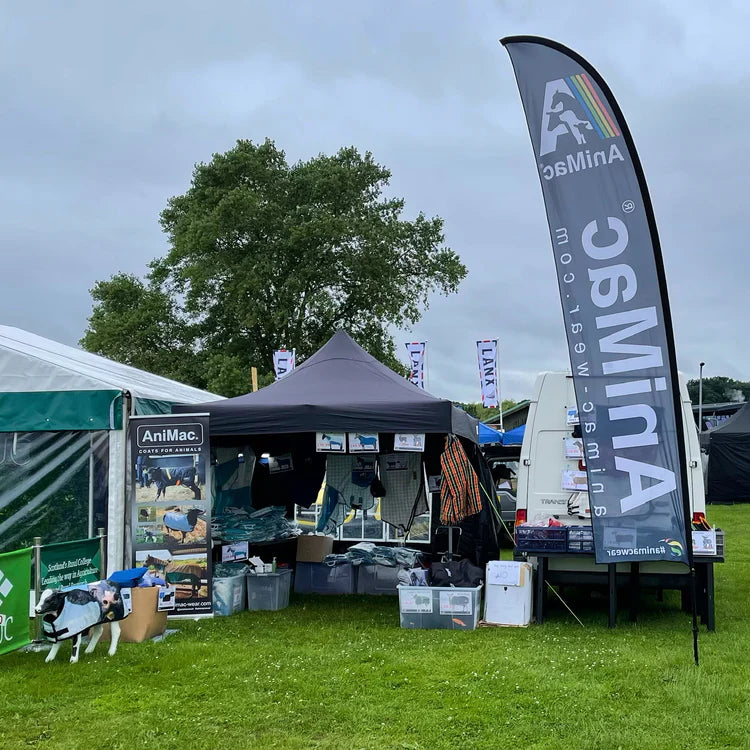 Outdoor AniMac booth at an event, with a black tent, banners, product displays, and a table with bins. A model cow and promotional signs are visible on grass under cloudy skies.