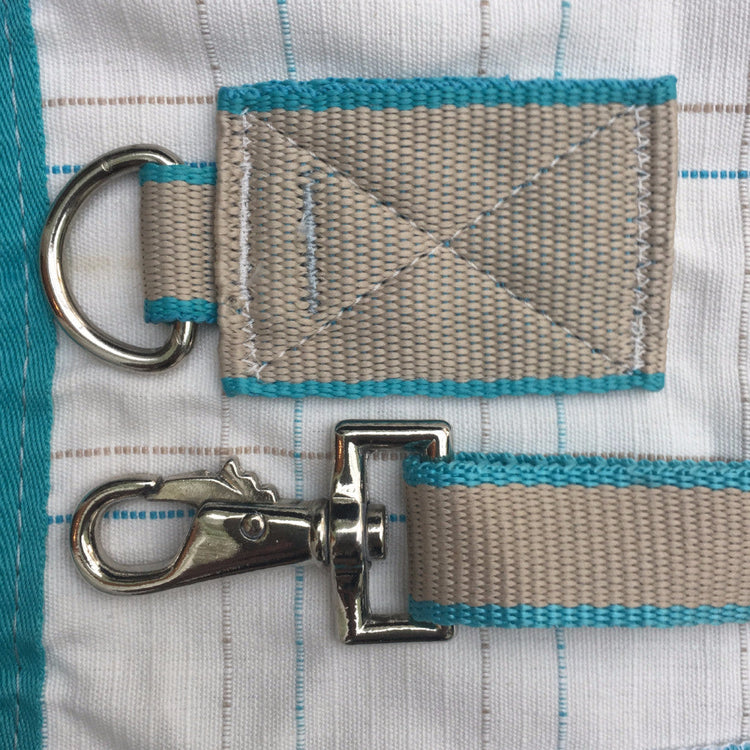 Close-up of the AniMac Allrounder Fly Rug Combo, highlighting beige and turquoise stitching, a metal D-ring, and swivel snap hook on webbing straps, set against a striped background.