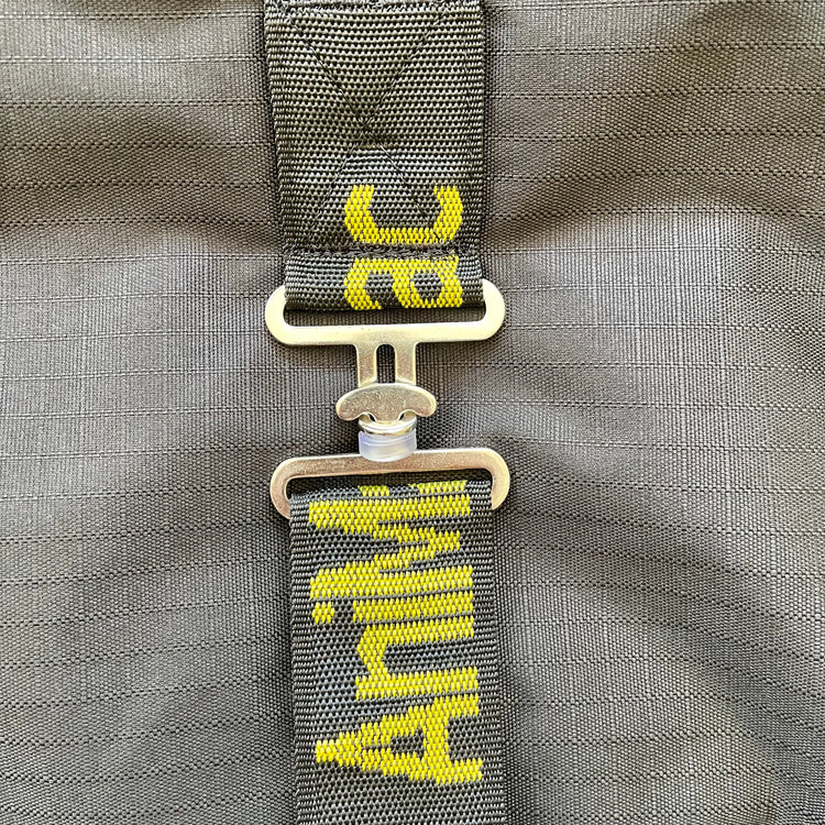 Close-up of the adjustable belly strap with yellow text, fastened by a metallic buckle on a gray, breathable Ram Rug | Waterproof. The strap shows partial text "Anim" and "ac" against the fabric background.
