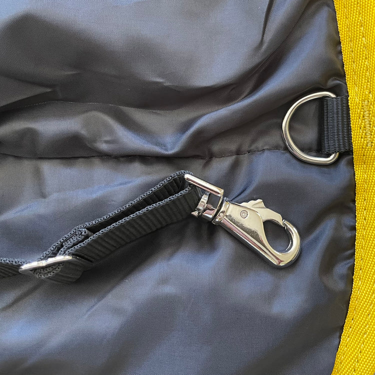 Close-up of the metal clasp hook and D-ring on a black strap, sewn onto dark fabric with yellow trim—features of the Ram Rug | Waterproof, designed for durability and protection.