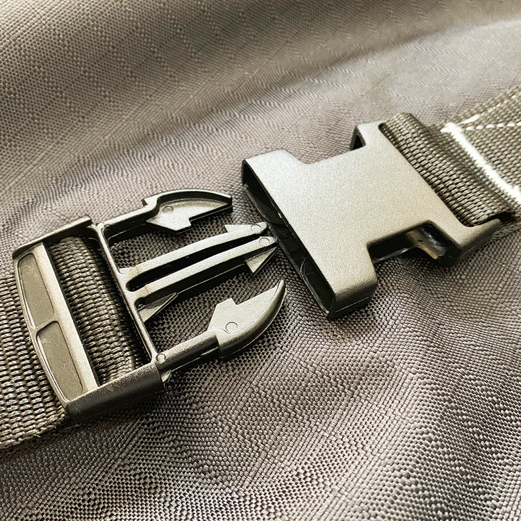 A close-up of a black side-release plastic buckle on a gray fabric strap from the Super Calf Jacket | Waterproof, displayed on a textured gray surface. The partially undone buckle reveals its prongs and clasp mechanism.
