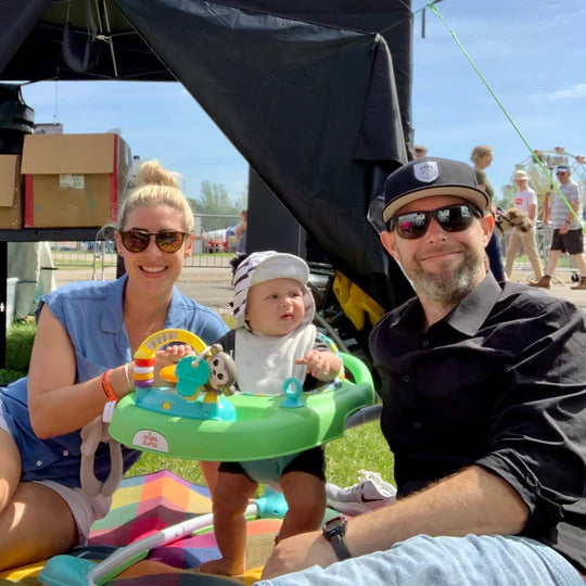 A smiling woman, a baby in a green activity center, and a bearded man in sunglasses and a cap pose together outdoors on a sunny day, sitting on a blanket at what appears to be a festival or fair.